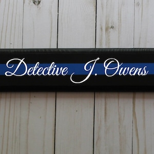 May include: Black wooden sign with a blue stripe and white text that reads "Detective J. Owens".