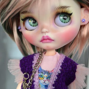 Può includere: Bambola con occhi verdi, fard rosa e labbra rosa. La bambola indossa un gilet viola sopra una maglietta rosa con una grafica di gatto. Una catena d'argento e orecchini a fiore completano il look. La bambola ha capelli rosa e verdi.