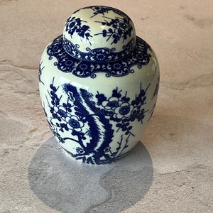 May include: A small, round, lidded ceramic jar with a cream base and detailed blue floral patterns. The jar features a floral design on the body and lid, creating a decorative piece. The jar is likely used for storage or display.