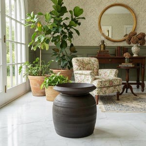 May include: A dark brown, cylindrical ceramic stool or planter with a wide, flared rim and horizontal ribbed detailing along the body. The piece has a matte finish and sits on a white marble floor in front of several potted green plants.