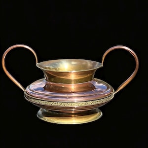 May include: A two-handled copper and brass bowl featuring a wide, polished copper body with a central band of intricate, embossed gold-toned metal detailing. The vessel includes a flared brass rim, a circular brass base, and two curved handles.