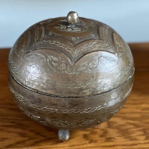 May include: A spherical brass container with a removable lid, featuring intricate engraved patterns and a small central knob. The object is supported by three small, rounded feet and displays a weathered, metallic finish with visible etched details.