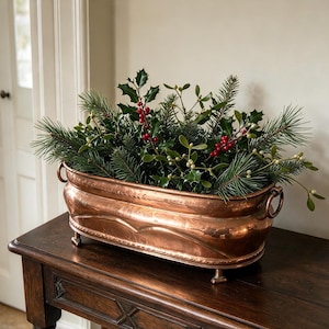 May include: An oval copper planter with a polished finish, featuring two side ring handles and four small feet. The planter is filled with a dense arrangement of green pine needles, holly leaves with bright red berries, and mistletoe sprigs.
