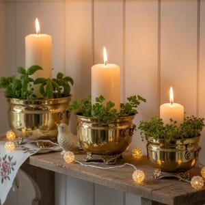 May include: Three brass pots with feet and side handles, each containing a green plant and a tall white pillar candle. The candles are lit, emitting a warm glow. A string of small, glowing spherical lights is draped in front of the pots.