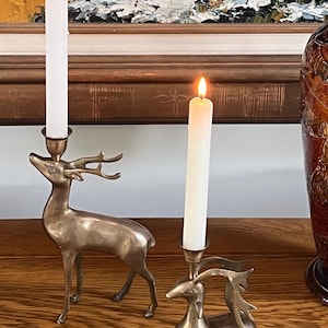 May include: Two brass deer-shaped candle holders, one standing and one lying down, each featuring a tall, slender white taper candle with a lit flame. The metallic figures have smooth, polished surfaces and detailed antlers holding the candles.