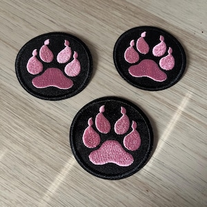 May include: Three circular black patches featuring a pink paw print design. The paw prints are embroidered in a light pink colour. The patches are on a light brown wooden surface.