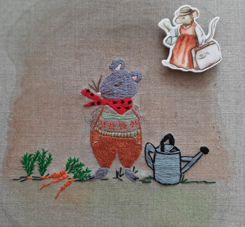 K&ouml;nnte beinhalten: Gestickte Abbildung einer grauen Maus, die einen rot-wei&szlig;en Schal und eine orangefarbene Hose tr&auml;gt und eine Gie&szlig;kanne h&auml;lt. Die Maus steht neben einem St&uuml;ck gestickter gr&uuml;ner Karotten.
