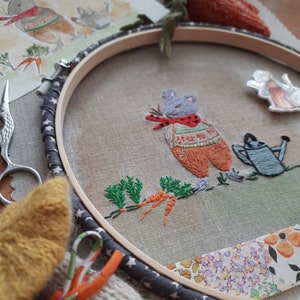 K&ouml;nnte beinhalten: Stickerei in einem Stickrahmen mit einer grauen Maus, die einen rot-orangenen Pullover tr&auml;gt und eine Karotte h&auml;lt. Die Maus ist von gr&uuml;nen Bl&auml;ttern und Karotten umgeben. Der Rahmen ist grau mit wei&szlig;en Sternen und befindet sich auf einem Stoffhintergrund mit einem Blumenmuster.