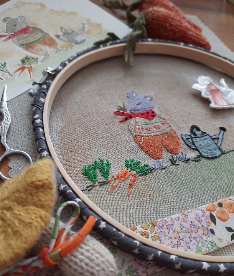 K&ouml;nnte beinhalten: Stickerei in einem Stickrahmen mit einer grauen Maus, die einen rot-orangenen Pullover tr&auml;gt und eine Karotte h&auml;lt. Die Maus ist von gr&uuml;nen Bl&auml;ttern und Karotten umgeben. Der Rahmen ist grau mit wei&szlig;en Sternen und befindet sich auf einem Stoffhintergrund mit einem Blumenmuster.
