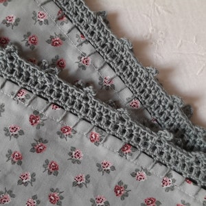 Può includere: Primo piano di un tessuto grigio con un piccolo motivo floreale rosa. Il tessuto è rifinito con due strati di pizzo all'uncinetto grigio.