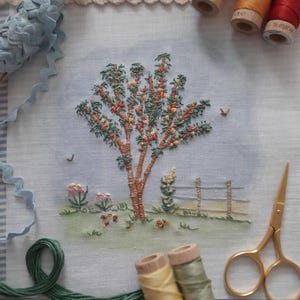 Peut inclure: Panneau de lin brodé représentant un arbre en fleurs détaillé, avec une clôture et des fleurs dans un jardin. Le panneau est entouré de fils à broder de différentes couleurs.