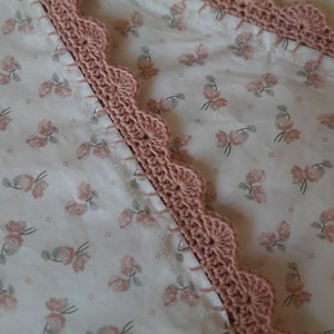 Può includere: Primo piano di un bordo in crochet rosa cucito su un tessuto bianco con un motivo floreale. Il bordo ha un bordo a festoni ed è realizzato con un punto a crochet semplice.