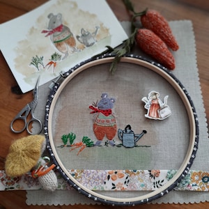 K&ouml;nnte beinhalten: Stickerei auf einem Stickrahmen mit einer grauen Maus mit einem rot-wei&szlig;en Pullover, die eine Gie&szlig;kanne h&auml;lt, einer grauen Maus mit einem roten Kleid und einem Korb und einer gr&uuml;nen Pflanze mit Karotten. Der Rahmen ist mit einem Blumenband verziert.