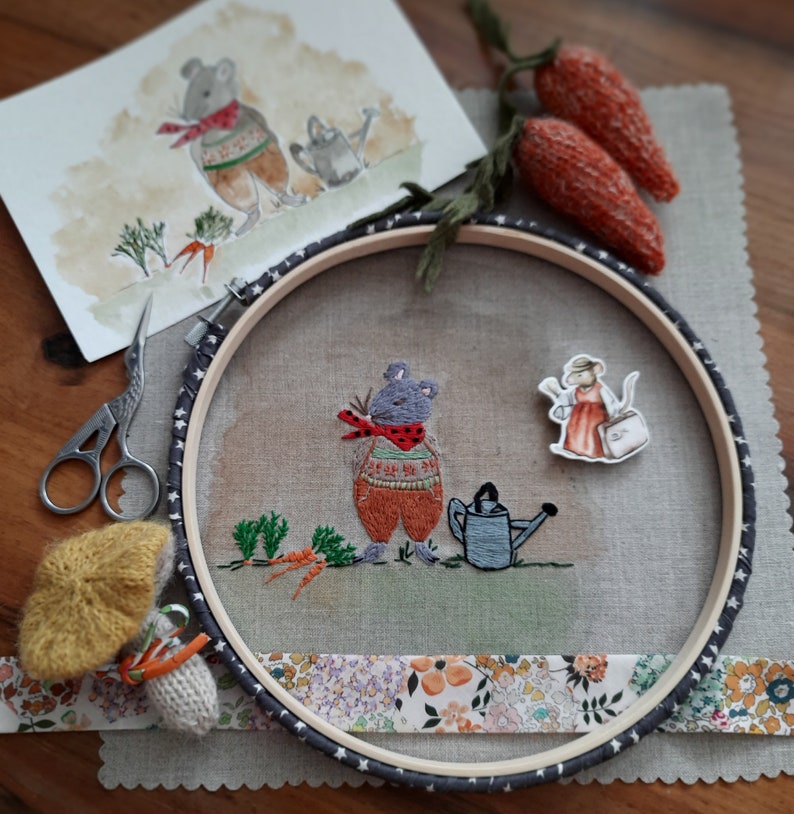 K&ouml;nnte beinhalten: Stickerei auf einem Stickrahmen mit einer grauen Maus mit einem rot-wei&szlig;en Pullover, die eine Gie&szlig;kanne h&auml;lt, einer grauen Maus mit einem roten Kleid und einem Korb und einer gr&uuml;nen Pflanze mit Karotten. Der Rahmen ist mit einem Blumenband verziert.