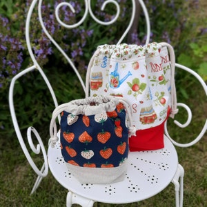 May include: Two fabric drawstring bags. One bag is white with a red, green, and blue floral pattern and the text "Garden Party". The other bag is navy blue with a white and red strawberry pattern.