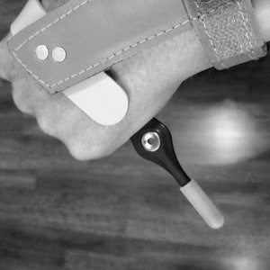 Può includere: Un'immagine in bianco e nero di una mano che tiene uno strumento bianco e nero con una punta bianca. Lo strumento ha un piccolo anello di metallo nella parte superiore.