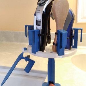 May include: A blue and white plastic holder with a clamp base, holding a hair clipper, a round brush, a wooden brush, and a blue hair styling tool. The holder is attached to a white countertop.