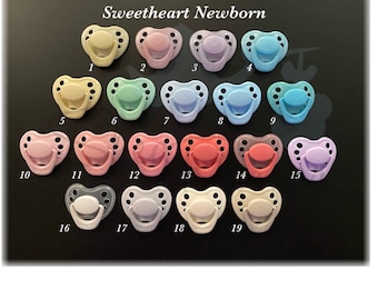 May include: Nineteen pacifiers in various colors, including pink, blue, green, and purple, arranged in rows on a black surface. The pacifiers are labeled with numbers 1 through 19.