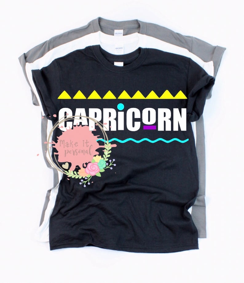 Op de afbeelding: Zwart t-shirt met het woord "CAPRICORN" in het wit, boven een geel driehoekontwerp en een turquoise golf. Het t-shirt is gelaagd met een wit en grijs t-shirt. Het ontwerp heeft een dierenriemthema.