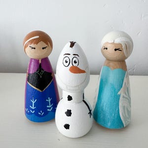 May include: Three hand-painted wooden peg dolls. The left doll has brown hair, a purple cape, and a blue dress with snowflake patterns. The centre doll is a white snowman with a carrot nose and black buttons. The right doll has white hair and a light blue dress.