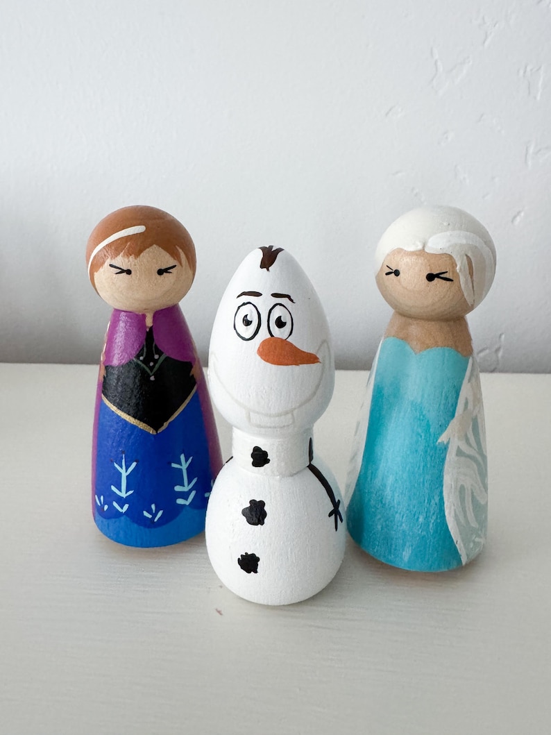 May include: Three hand-painted wooden peg dolls. The left doll has brown hair, a purple cape, and a blue dress with snowflake patterns. The centre doll is a white snowman with a carrot nose and black buttons. The right doll has white hair and a light blue dress.