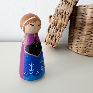 May include: A wooden peg doll painted to resemble a character with brown hair, a black bodice, and a blue skirt featuring light blue floral patterns. The figure has a purple cape painted on the back and sides, with a simple face and white headband.