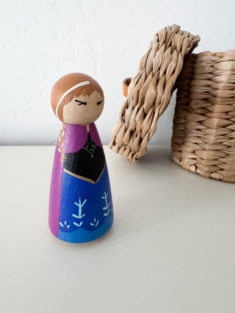 May include: A wooden peg doll painted to resemble a character with brown hair, a black bodice, and a blue skirt featuring light blue floral patterns. The figure has a purple cape painted on the back and sides, with a simple face and white headband.