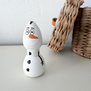 May include: A white wooden peg doll shaped like a snowman with a painted orange carrot nose, black coal buttons, a brown twig arm, and a brown hair tuft on top. The figure features a wide, smiling expression with large eyes and eyebrows.