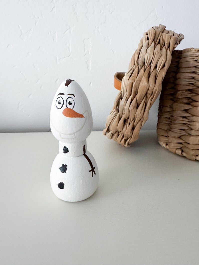 May include: A white wooden peg doll shaped like a snowman with a painted orange carrot nose, black coal buttons, a brown twig arm, and a brown hair tuft on top. The figure features a wide, smiling expression with large eyes and eyebrows.