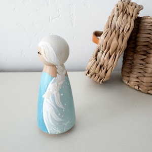 May include: A wooden peg doll painted with a light blue dress featuring white snowflake details and a long white braided hairstyle. The figure has a smooth, rounded head with simple painted facial features and a natural wood finish on the neck.