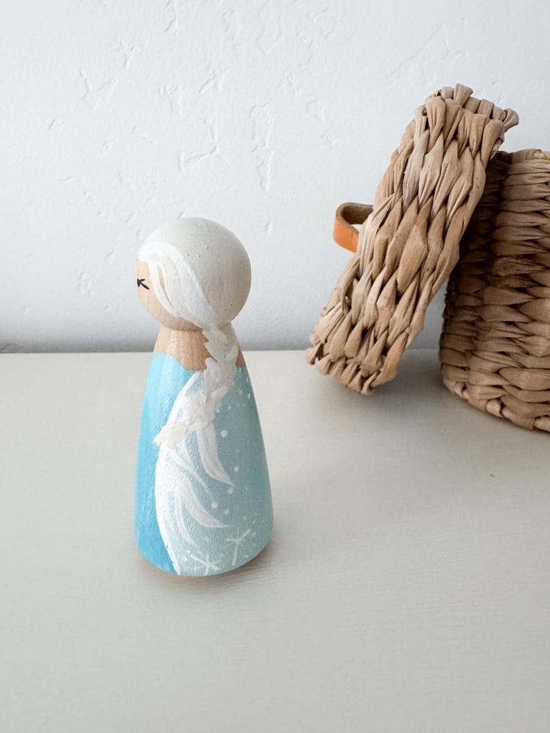 May include: A wooden peg doll painted with a light blue dress featuring white snowflake details and a long white braided hairstyle. The figure has a smooth, rounded head with simple painted facial features and a natural wood finish on the neck.