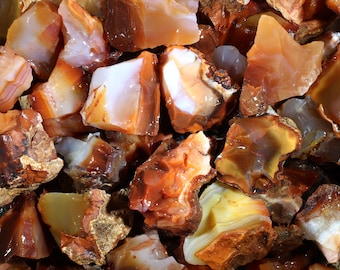 May include: A collection of rough, natural carnelian stones. The stones are a mix of orange, red, and brown colors with white banding. The carnelian is a semi-precious gemstone.