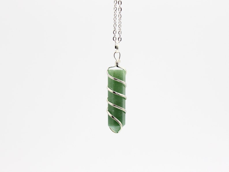 May include: A green gemstone pendant necklace. The pendant is a cylindrical shape, wrapped with a silver-colored wire in a spiral design. The necklace chain is also silver-colored and hangs against a white background.