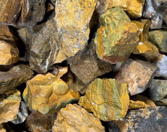 May include: A collection of raw, unpolished stones featuring a variety of earthy tones including mustard yellow, olive green, deep brown, and gray. The rocks exhibit rough, jagged textures with visible mineral layering and natural surface variations.