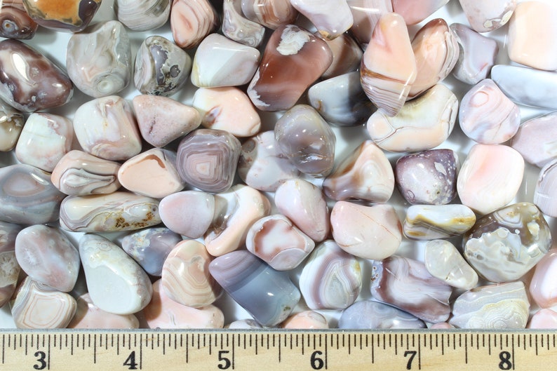 May include: A collection of tumbled agate stones in shades of pink, white, and gray. The stones are smooth and polished, with intricate banding patterns. The stones are arranged on a wooden ruler, with the measurements 3 to 8 inches marked.