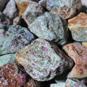 May include: A close-up of a group of rough, green, and brown stones with pink flecks. The stones are tumbled and have a natural, earthy look.
