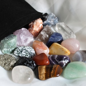 May include: A collection of tumbled stones in a variety of colors and textures, including amethyst, rose quartz, tiger's eye, and clear quartz. The stones are arranged in a loose pile on a white fabric background.