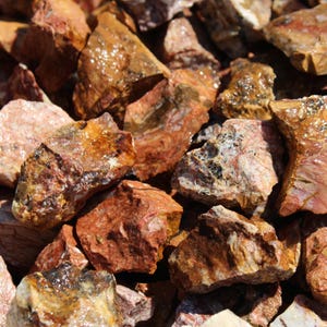 May include: Close-up of a pile of natural, rough-cut rocks. The rocks vary in size and shape, with a mix of reddish-brown, tan, and pink hues. Some rocks have a wet, glistening appearance.