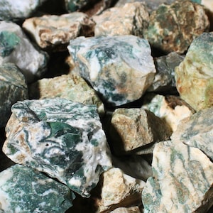 May include: A collection of raw, unpolished moss agate stones featuring a mix of deep forest green, translucent white, and earthy tan inclusions. These rough mineral specimens display irregular, jagged shapes with varied textures and natural patterns.