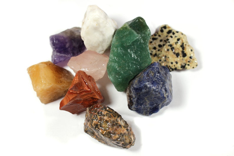 May include: A collection of raw gemstones in various colors, including purple amethyst, white quartz, green aventurine, and speckled dalmatian jasper. Other stones are shades of red, yellow, pink, and blue. The stones are arranged on a white surface.