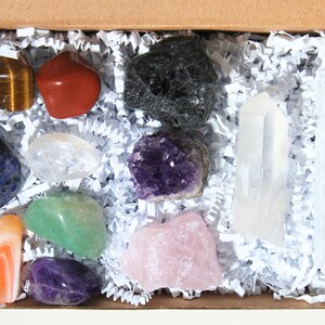 May include: A box filled with white crinkle paper containing a variety of crystals and stones, including amethyst, rose quartz, clear quartz, tiger's eye, red jasper, green aventurine, lapis lazuli, and black obsidian.
