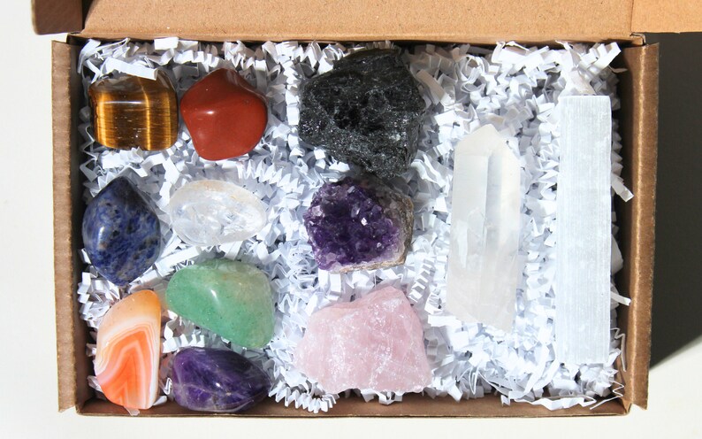 May include: A box filled with white crinkle paper containing a variety of crystals and stones, including amethyst, rose quartz, clear quartz, tiger's eye, red jasper, green aventurine, lapis lazuli, and black obsidian.