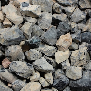 May include: A pile of various-sized rocks in shades of gray, tan, and black. The rocks have rough, textured surfaces and irregular shapes. The image shows a close-up view of the rocks, filling the frame.