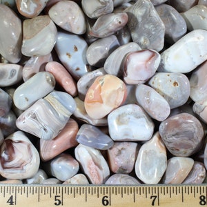 May include: A collection of tumbled agate stones in shades of gray, white, and pink. The stones are smooth and polished, with intricate patterns of banding and swirls. The stones are arranged on a wooden ruler, with the measurements 3 to 9 inches marked.