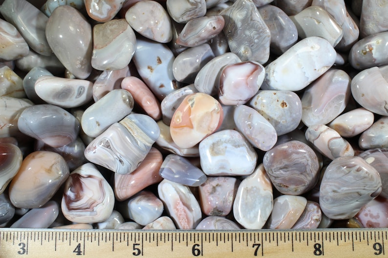 May include: A collection of tumbled agate stones in shades of gray, white, and pink. The stones are smooth and polished, with intricate patterns of banding and swirls. The stones are arranged on a wooden ruler, with the measurements 3 to 9 inches marked.