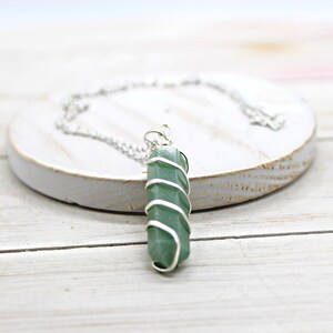 May include: A green crystal pendant wrapped in silver-colored wire, suspended from a delicate silver chain. The necklace rests on a white, circular, wooden surface. The crystal is a vibrant green color.