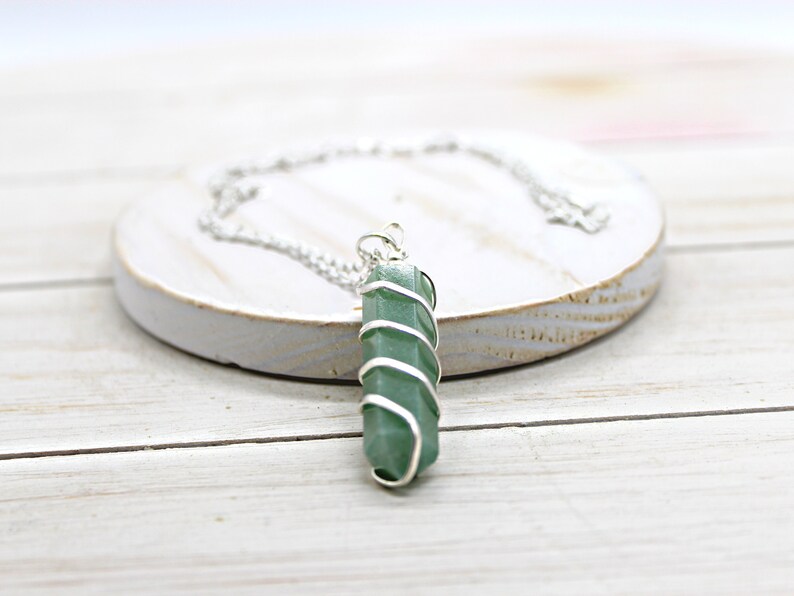 May include: A green crystal pendant wrapped in silver-colored wire, suspended from a delicate silver chain. The necklace rests on a white, circular, wooden surface. The crystal is a vibrant green color.