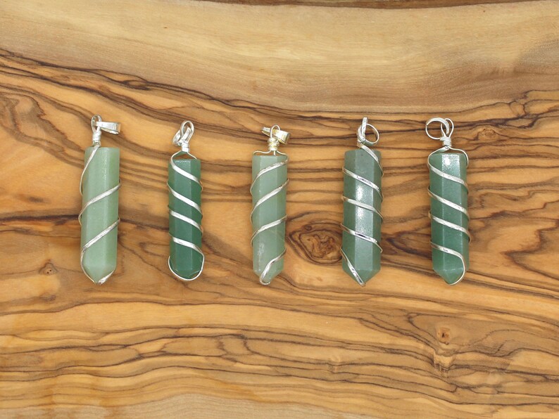 May include: Six green gemstone pendants with silver wire wrapping are displayed on a wooden surface. Each pendant has a hexagonal shape and a silver loop at the top for hanging. The pendants vary slightly in shade of green.