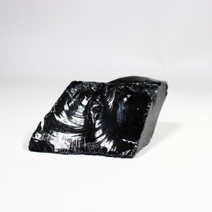 Puede incluir: Un trozo en bruto, de forma irregular, de obsidiana, un vidrio volc&aacute;nico, con una superficie brillante y reflectante. La piedra negra tiene una apariencia fracturada, que atrapa la luz y crea reflejos. El fondo es blanco.
