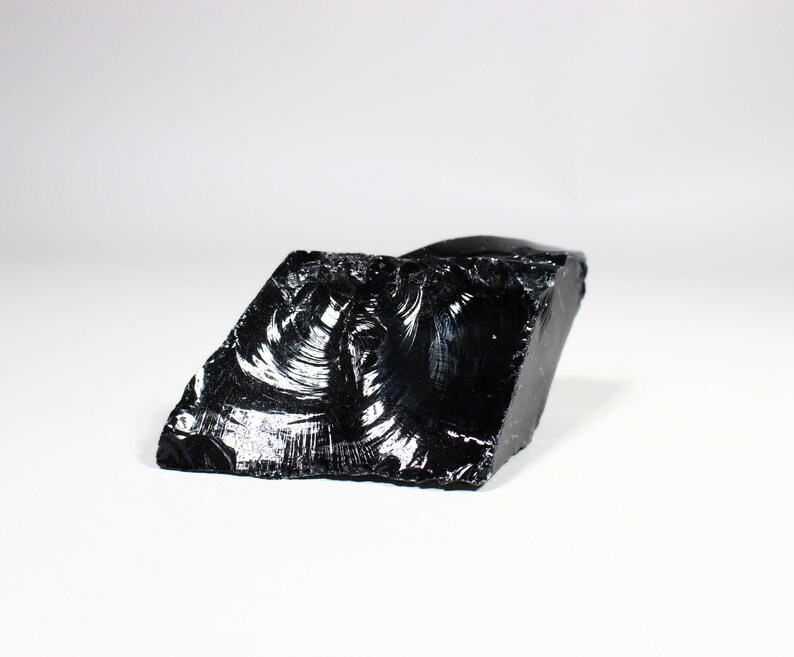 Puede incluir: Un trozo en bruto, de forma irregular, de obsidiana, un vidrio volc&aacute;nico, con una superficie brillante y reflectante. La piedra negra tiene una apariencia fracturada, que atrapa la luz y crea reflejos. El fondo es blanco.